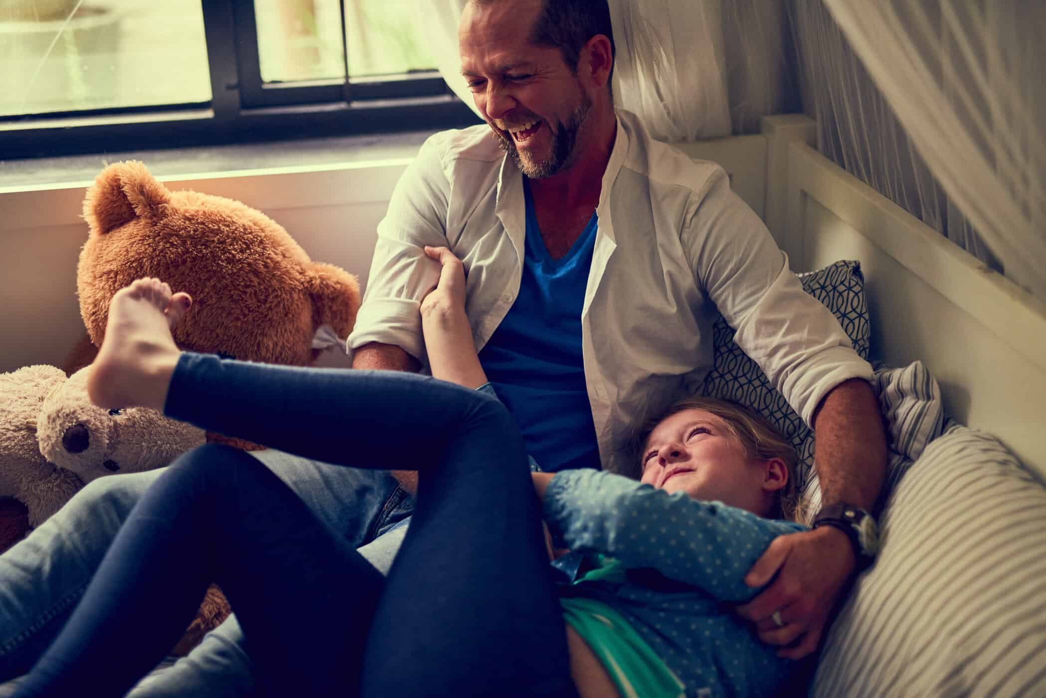 ADHS bei Kindern – Vater spielt lachend mit seiner Tochter auf dem Sofa, umgeben von Teddybären.