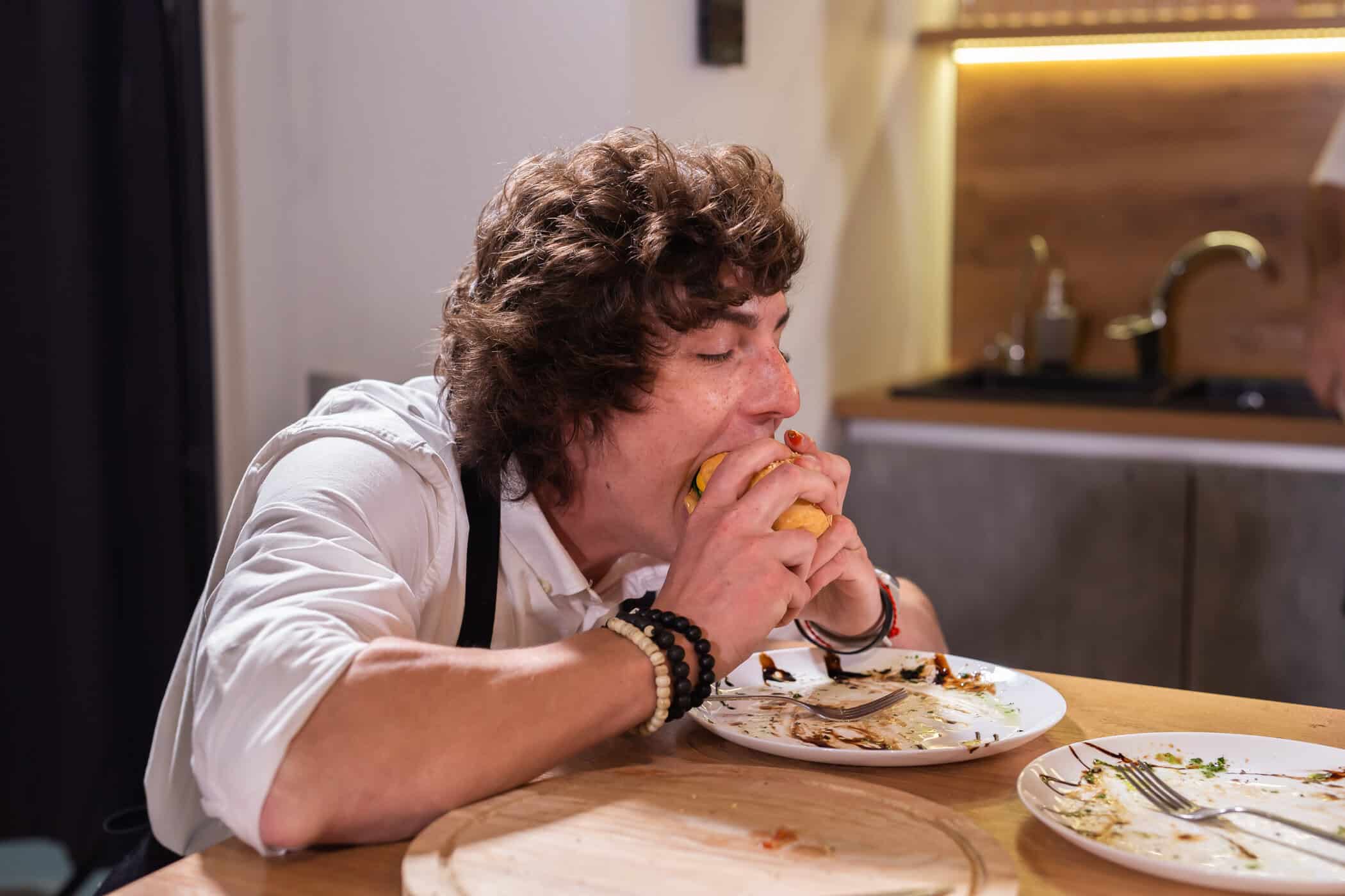 Junger Mann isst hastig mit beiden Händen an einem Tisch mit leeren Tellern symbolisiert ADHS und Binge-Eating.