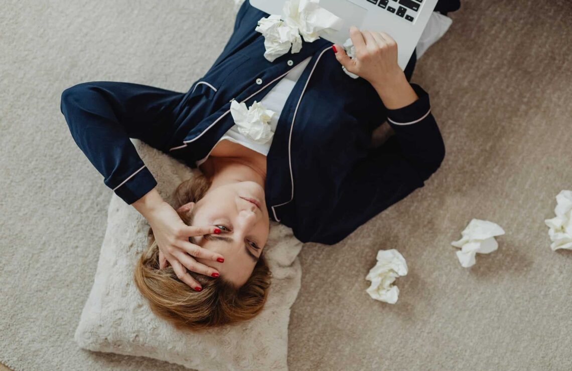 kranke Frau liegt auf dem rücken am Boden, laptop auf dem Bauch, Taschentücher liegen um sie herum