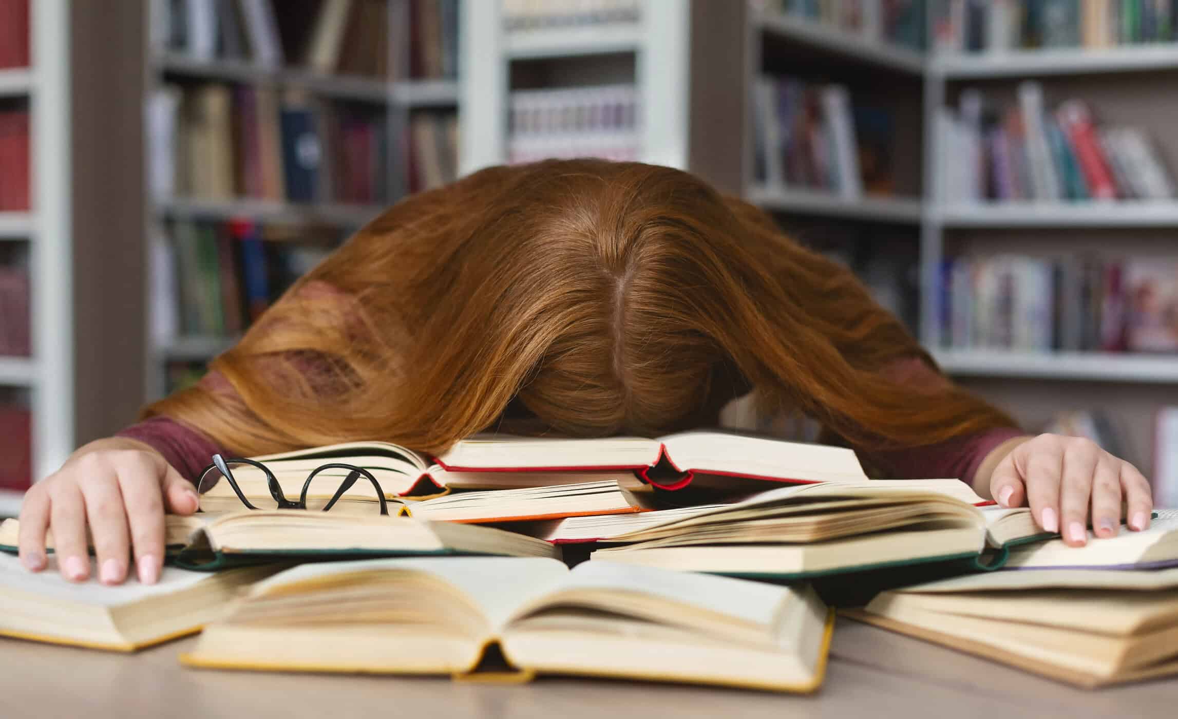 Erschöpfte Frau mit dem Kopf auf einem Stapel aufgeschlagener Bücher in einer Bibliothek symbolisiert Lernen mit ADHS.