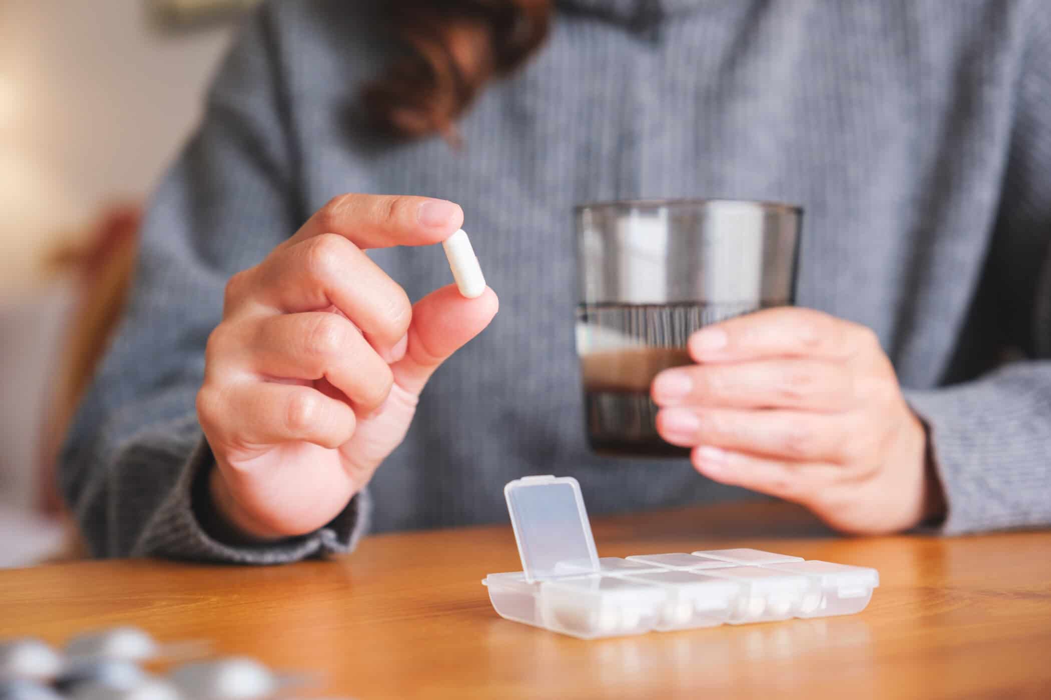 Person h&auml;lt eine Tablette &uuml;ber einer ge&ouml;ffneten Pillenbox, daneben ein Glas Wasser &ndash; sinnbildlich f&uuml;r die t&auml;gliche Einnahme und die Medikinet Erfahrungen Betroffener im Alltag