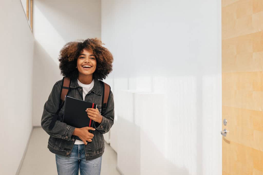 Lächelnde Studentin mit Rucksack und Notizblock im Flur symbolisiert gelingendes studieren mit ADHS.