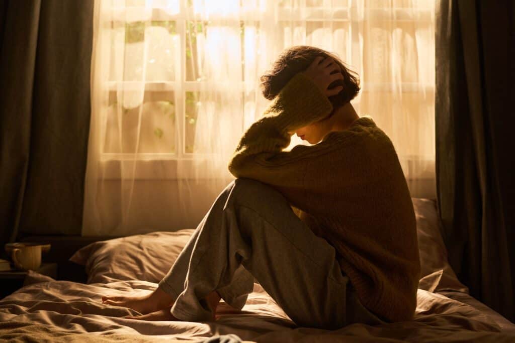 Auf dem Bett sitzende Frau hält sich den Kopf und symbolisiert Anstrengung durch innere Unruhe