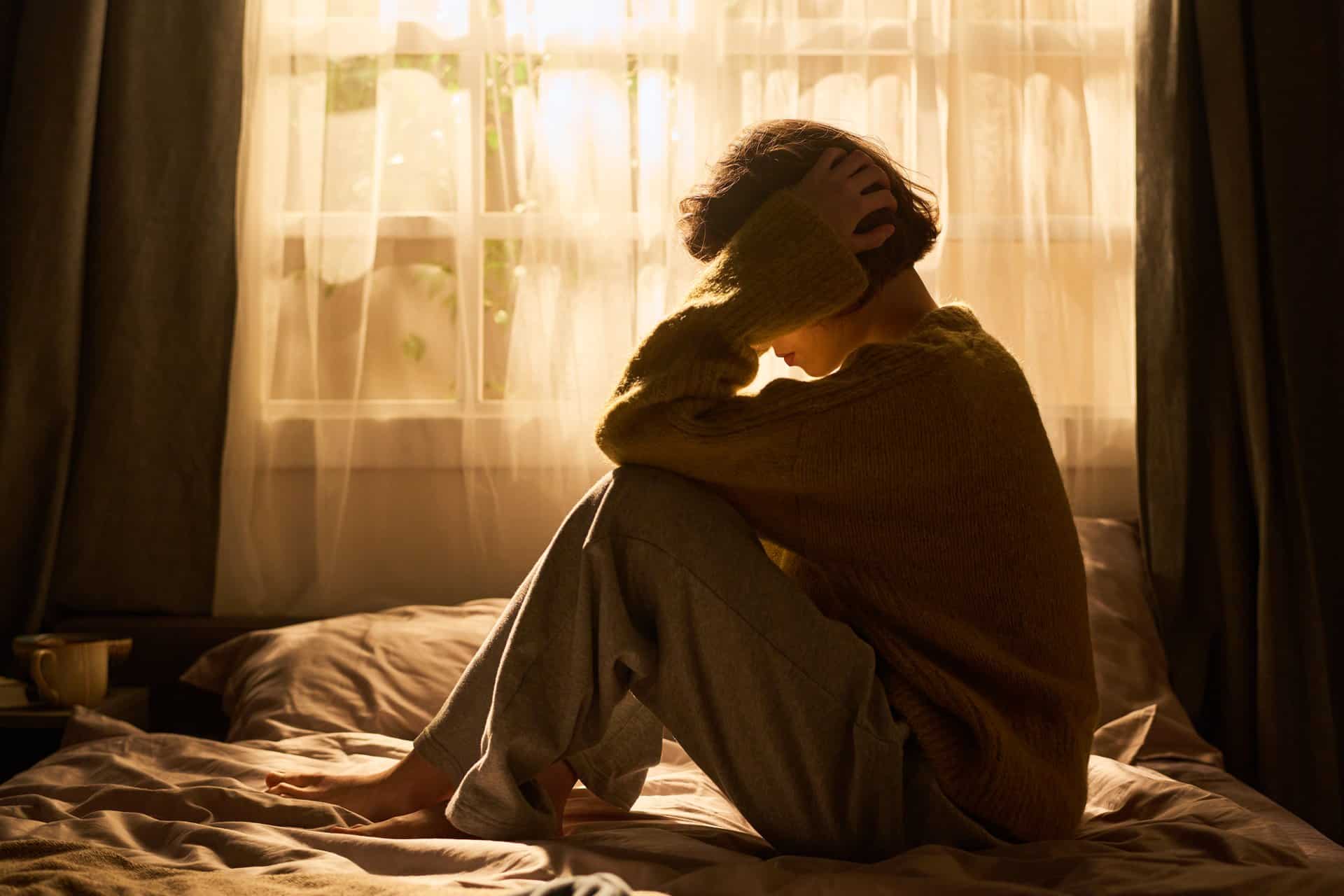 Auf dem Bett sitzende Frau h&auml;lt sich den Kopf und symbolisiert Anstrengung durch innere Unruhe